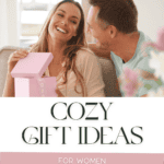 A man and woman sit next to each other smiling as she opens a pink box with the words across saying cozy gift ideas for women