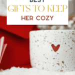 A speckled ceramic mug with a heart design sits on a soft white surface. Above it, text reads "The Bargain Sisters Best Gifts to Keep Her Cozy" in various fonts and colors, while a red and pink background complements the cozy vibe—a perfect pairing with cool baking gadgets for warmth.