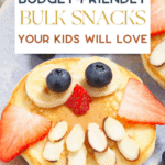 A playful pancake topped with banana slices, blueberries, a strawberry slice, and almond slivers to resemble a smiling face. Text reads: "The Bargain Sisters: Budget-Friendly Bulk Snacks Your Kids Will Love," crafted using our cool baking gadgets for extra fun!.