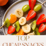 A plate showcasing skewered fruit slices—strawberries, bananas, grapes, oranges, and kiwis. An orange sits beside it. Text overlay reads, "Top Cheap Snacks to Buy in Bulk for Families" by The Bargain Sisters. Discover cool baking gadgets for crafting delightful treats at home!.