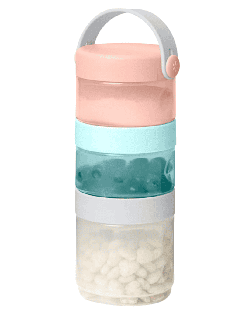 A stackable food container, perfect as travel gear for kids, features three compartments with assorted contents. The top lid is pink, while the middle section is mint green. Transparent sides reveal small items inside, and a grey handle on top ensures easy carrying.