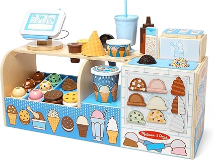 A wooden ice cream play set featuring cones, scoops, and toppings displayed on a blue and beige stand. It includes an order station with a "Monday Deals" menu board on the side. Various colorful ice cream scoops are arranged in compartments.