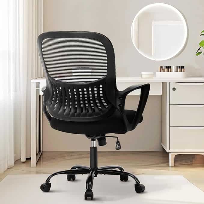 A black ergonomic office chair with a mesh back and armrests is positioned in front of a white desk, ready for those unbeatable Wednesday Deals. A round mirror is mounted on the wall above, and natural light filters through sheer white curtains from a nearby window.