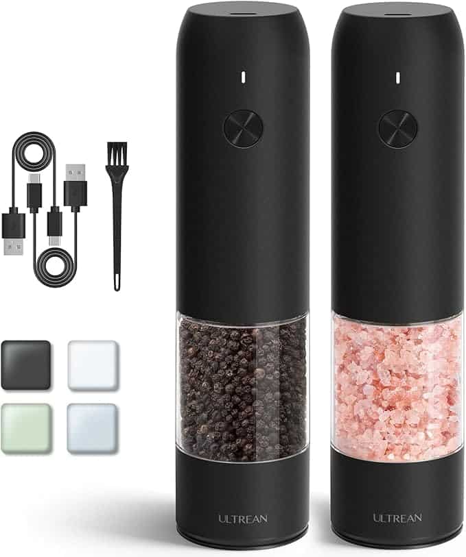 Two sleek black electric salt and pepper grinders feature transparent compartments filled with vibrant black peppercorns and pink Himalayan salt. Accompanied by two charging cables, a black cleaning brush, and four square white coasters, these are a perfect choice for Monday Deals. The brand name "Ultrean" is prominently displayed.