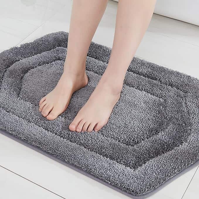 Bare feet standing on a soft, grey, rectangular bath mat with a textured, plush surface set on a white tiled floor, this comfort could be part of your Monday Deals.