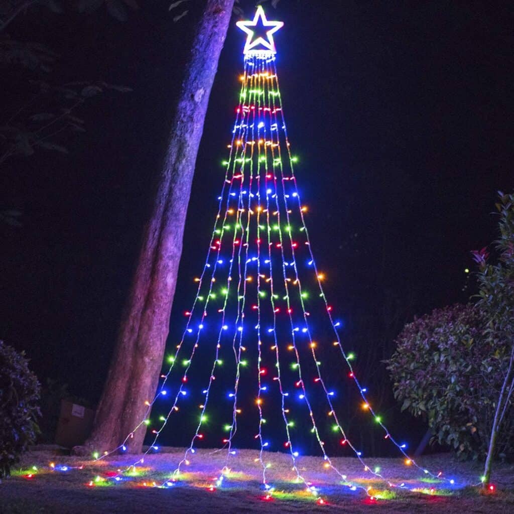 A festive outdoor display of vertical strings of colorful lights forms a lit Christmas tree with a glowing star on top. The dazzling arrangement, reminiscent of Thursday Deals excitement, drapes from a high point on a tree, creating a vibrant and cheerful scene against the night sky.