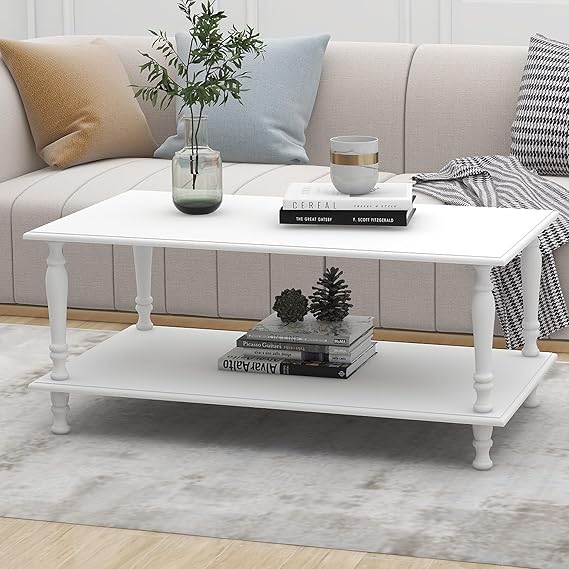 A white coffee table with a lower shelf showcases books and pinecones, offering an inviting touch. A glass vase with green foliage and a small round candle holder sit atop, perfectly capturing those cozy Monday Deal vibes. The table rests on a light rug beside a beige sofa adorned with patterned cushions.