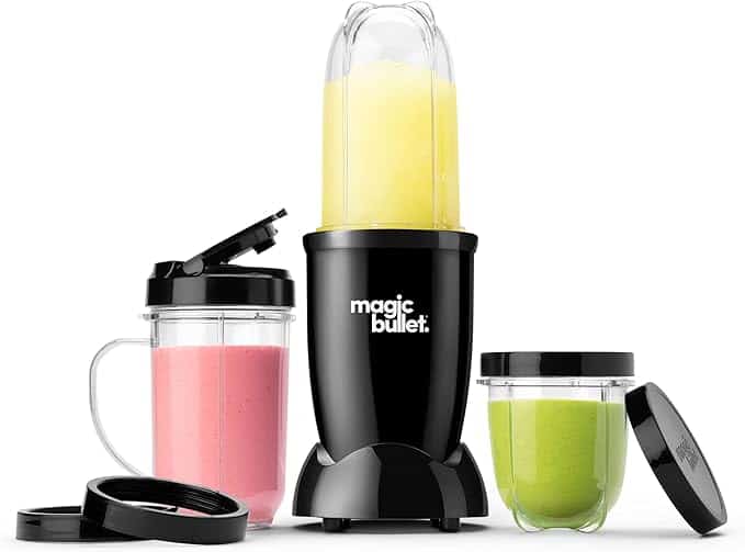 A black Magic Bullet blender is showcased alongside three cups of vibrant blended drinks, perfect for sharing during Friday Deals. The set features a tall cup with a yellow blend in the blender, a short cup with a pink drink and lid, and another short cup with a green drink and its lid beside it.