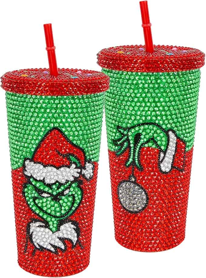 Two sparkling cups adorned with red and green rhinestones, perfect for Friday Deals. Each features a grinning character in a Santa hat and long green gloves, holding a shiny ornament on one cup. Both come with red lids and straws, making them an ideal festive bargain.