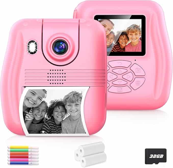 A pink camera with a digital screen displaying a group of smiling kids; beneath, a black-and-white photo is printed. Snap up this offer on Wednesday Deals! Includes four colored pens, extra photo paper rolls, and a 32GB memory card.