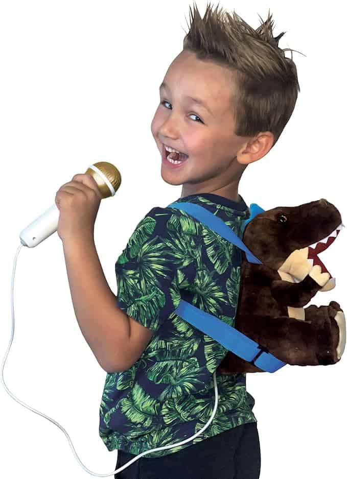 A smiling child with spiked hair holds a microphone and sports a brown plush dinosaur backpack, ready for Monday Deals. Wearing a green and black patterned shirt, the child faces away while cleverly glancing over their shoulder.