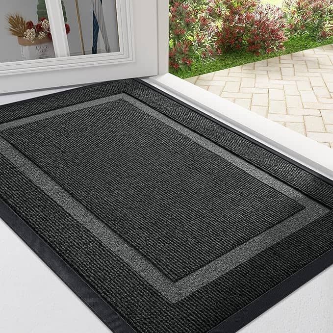 A charcoal gray doormat with a bordered design lies at an open door, hinting at cozy welcomes. Outside, Wednesday Deals await amidst a paved walkway and a garden bursting with blooming red and green foliage.