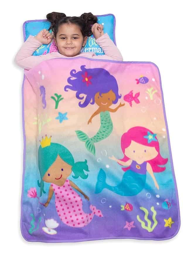 A smiling child lies on a vibrant blanket adorned with illustrations of three mermaids surrounded by sea creatures. The blanket's gradient background of blue, purple, and pink makes it a perfect find for Wednesday Deals shoppers seeking oceanic magic.