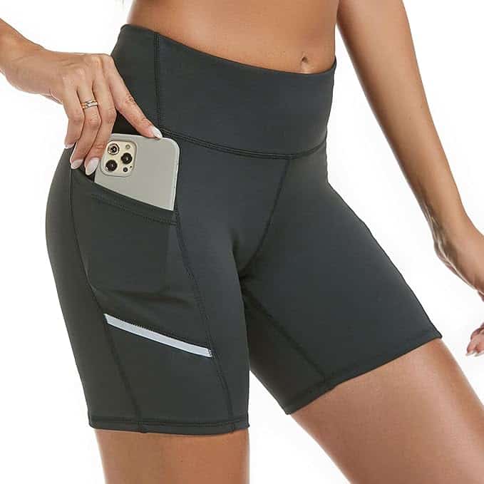 A person wearing high-waisted black workout shorts, perfect for those Wednesday Deals, is inserting a smartphone into the convenient pocket. The shorts feature a small reflective strip on the side, all set against a plain white background.