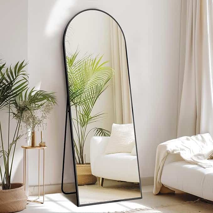 A modern bedroom corner showcases a tall, arched floor mirror with a black frame, reflecting a cozy white armchair and greenery. A beige blanket drapes over the armchair as natural light filters through large, white curtains—perfect for relaxing after shopping Tuesday Deals.