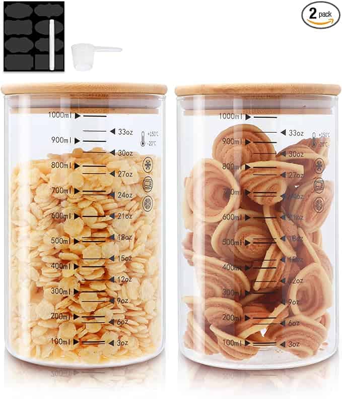 Two clear glass jars with wooden lids, perfect for cereal and cookies, feature measurement markings. A black sticker sheet and small scoop accompany the jars, which are available as a two-pack set. Snap up this stylish duo in our exclusive Thursday Deals!.