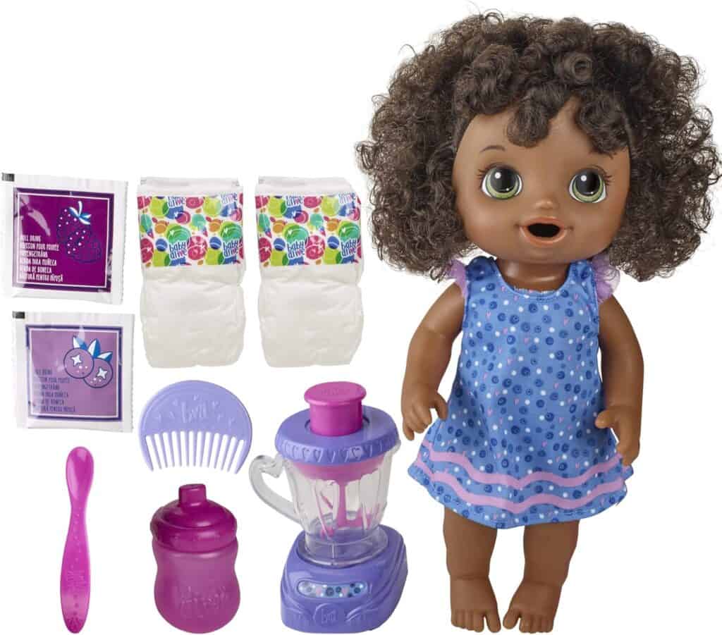 A doll with curly brown hair wearing a blue polka dot dress is surrounded by accessories: a purple and pink blender, a pink spoon, a purple sippy cup, a pink comb, two diapers, and two packets of doll food. Grab this delightful set in our exclusive Monday Deals!.