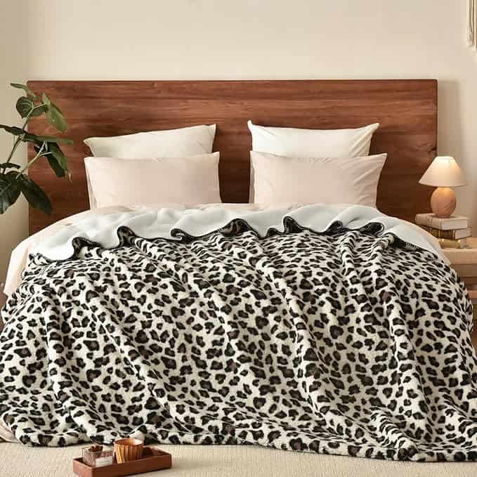 A cozy bedroom scene with a wooden headboard showcases a bed adorned with fluffy white pillows and a leopard print blanket. Perfect for Friday Deals, a small lamp and plant flank the bed, while a wooden tray with snacks invites relaxation.