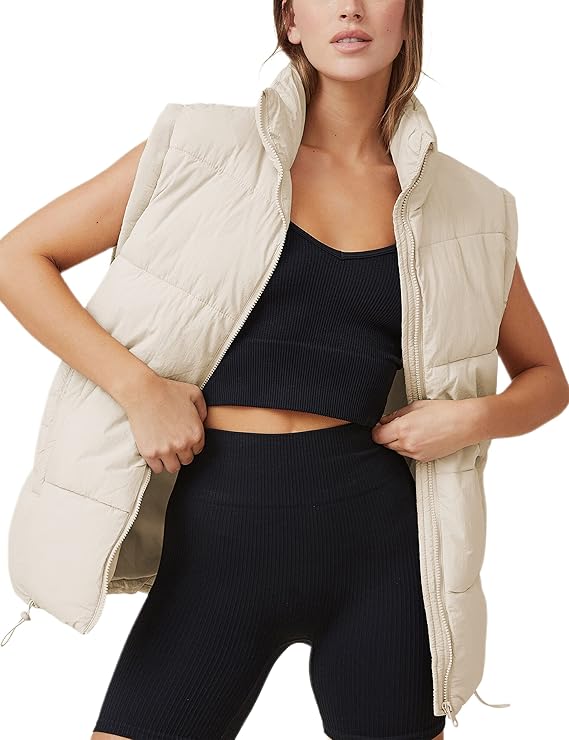 A woman showcases a beige puffer vest over a black ribbed sports bra and matching shorts, subtly adjusting the vest with her hands. It's an effortless look perfect for Thursday Deals, set against a simple backdrop.