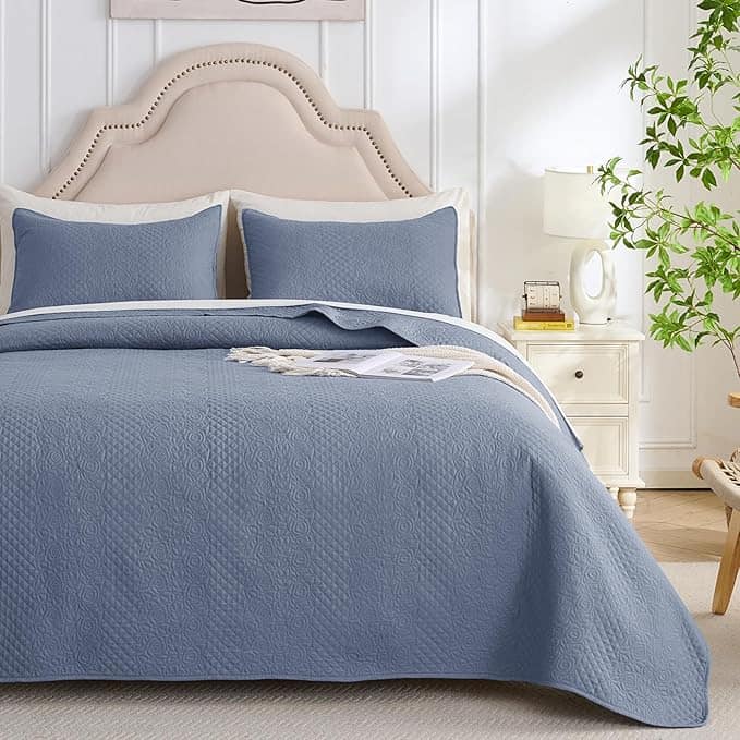 A neatly made bed with a light beige upholstered headboard and a blue quilted coverlet, flanked by two matching pillows. A white nightstand holds a lamp, a book, and a small plant. Discover stylish comfort with our Tuesday Deals, where even the nearby larger plant gets its moment against the white wall.