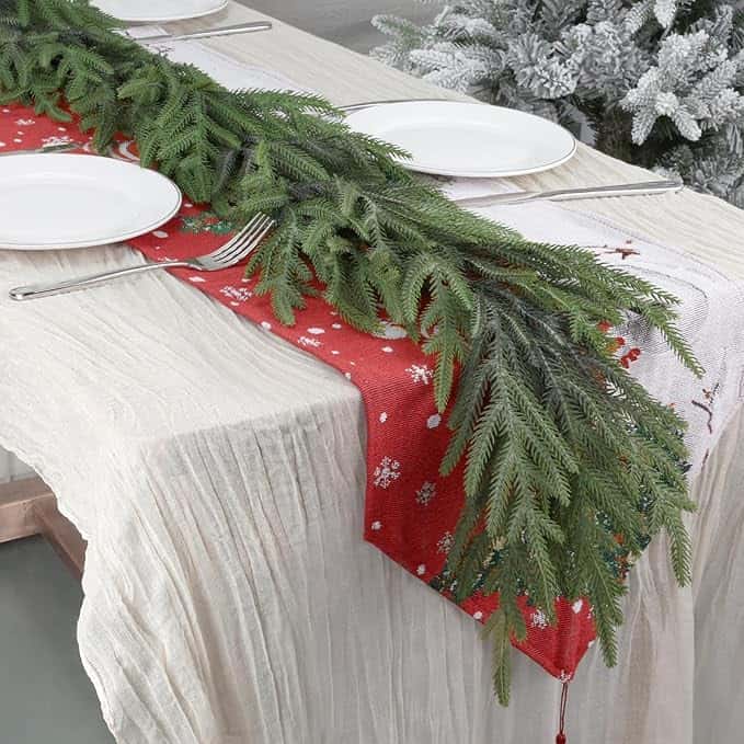 A festive table setting with white plates, silverware, and a red runner featuring snowflake patterns is ready for your Thursday Deals gathering. Green pine branches serve as a centerpiece, while a snowy tree graces the background, adding seasonal charm.