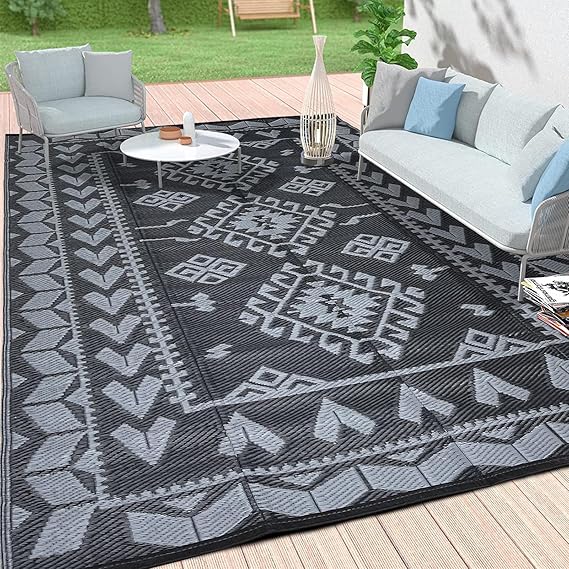 Outdoor patio with a black and gray geometric patterned rug on a wooden deck, perfect for those Monday Deals. Two cushioned chairs and a sofa flank a round white coffee table. A tall decorative lantern and green potted plant enhance the cozy setup, set against grass and a tree in the background.