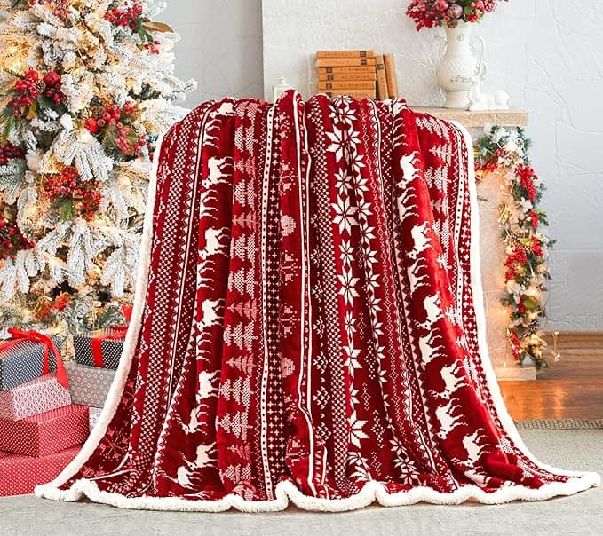 A red and white Christmas-themed blanket with reindeer and snowflake patterns is draped over a chair, set in a festive room with a decorated Christmas tree and wrapped presents. Don't miss the exclusive Wednesday Deals for this cozy holiday touch.