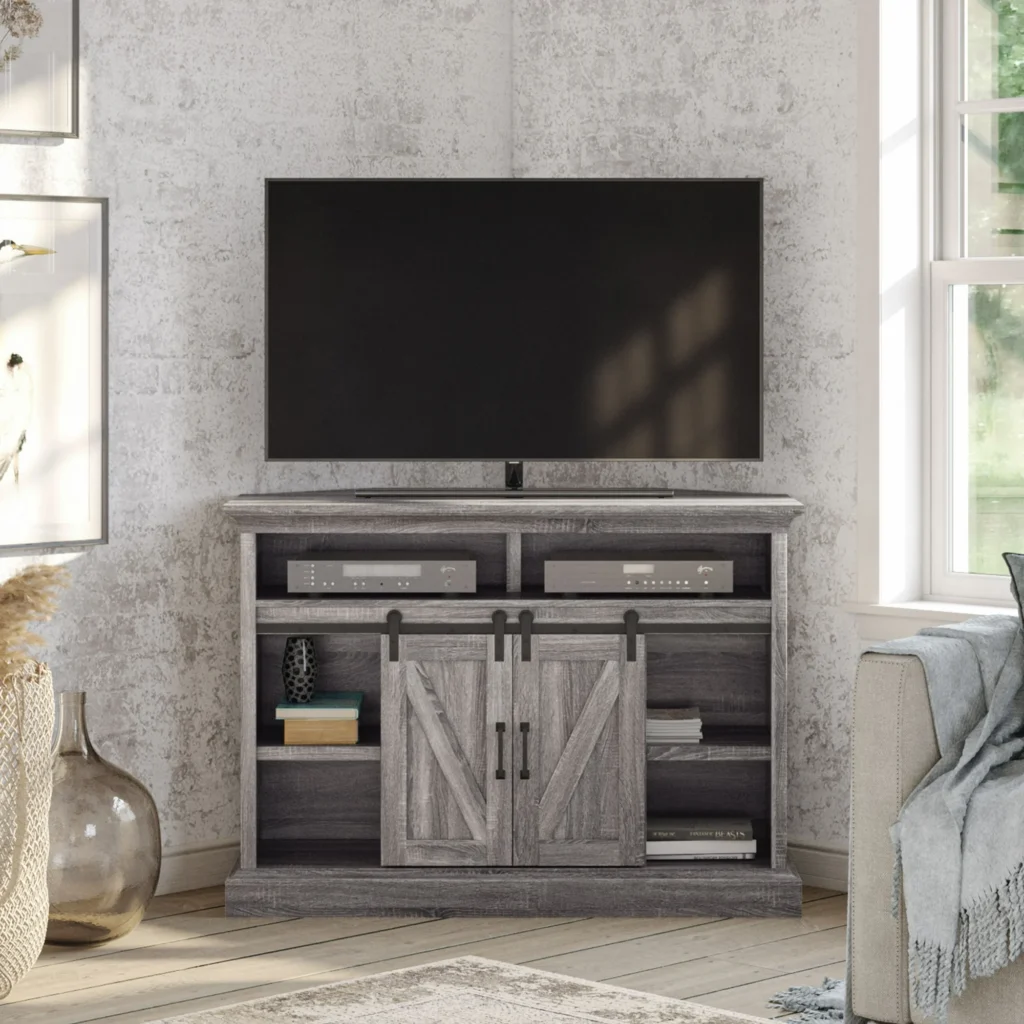 A rustic TV stand with a flat-screen TV sits atop, featuring barn-style sliding doors and open shelves adorned with decor items. Bathed in light from a large window, it's near a cozy chair draped with a blanket. Perfect for those Friday Deals, transforming your space into a stylish retreat.