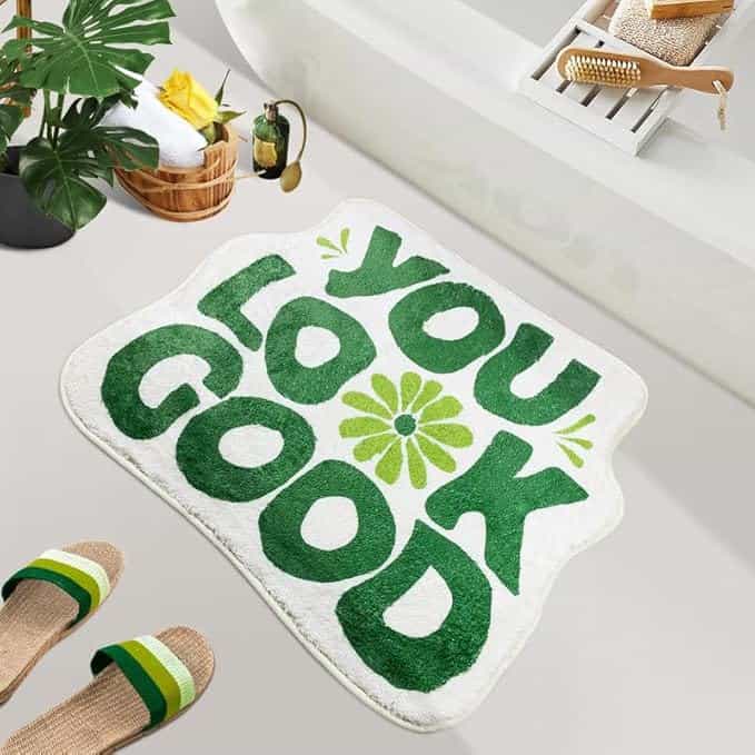 Bathroom scene featuring a white and green rug with "YOU LOOK GOOD" in bold letters. The area boasts a bathtub with bath accessories, a plant, a wicker basket with towels, and striped slippers on the floor—perfect ambiance for enjoying Thursday Deals relaxation.