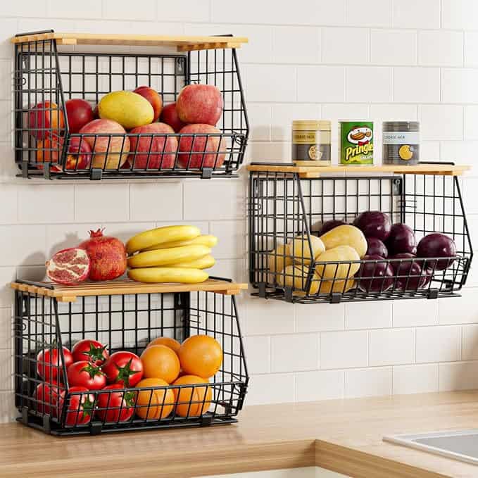 Wall-mounted black metal baskets offer a Tuesday Deals delight, holding apples, bananas, oranges, tomatoes, eggplants, and a pomegranate alongside three cans. These baskets rest on wood shelves against a white tiled wall above the kitchen counter.