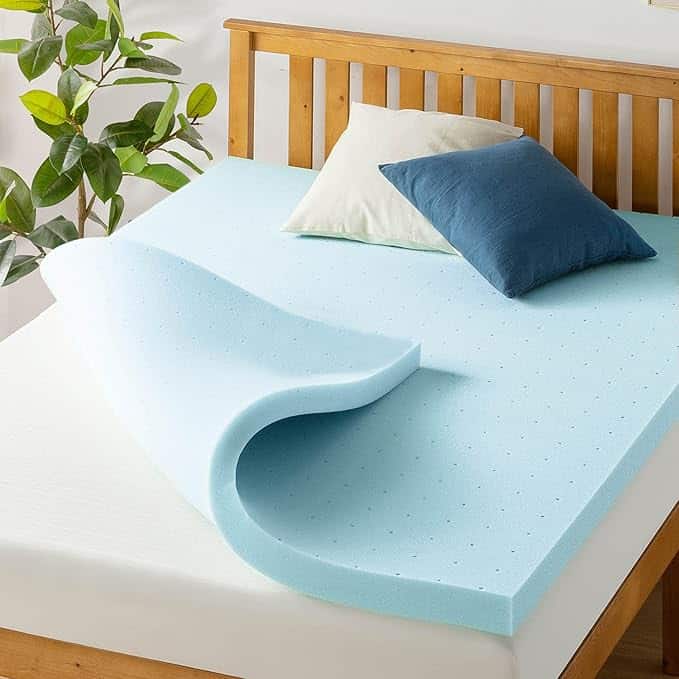 A memory foam mattress topper, light blue in color, is partially unfolded on a bed with a wooden headboard. Two pillows, one beige and one dark blue, rest near the headboard. A green potted plant is visible beside the bed, ready to bring comfort and style perfect for Wednesday Deals.