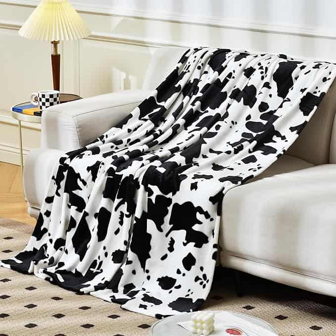 A soft, white blanket with black cow print draped over a beige sofa in the cozy living room sets the scene for Tuesday Deals. Beside the sofa, a table holds a cup and a stack of books. A lamp and patterned rug add warmth to this inviting space.