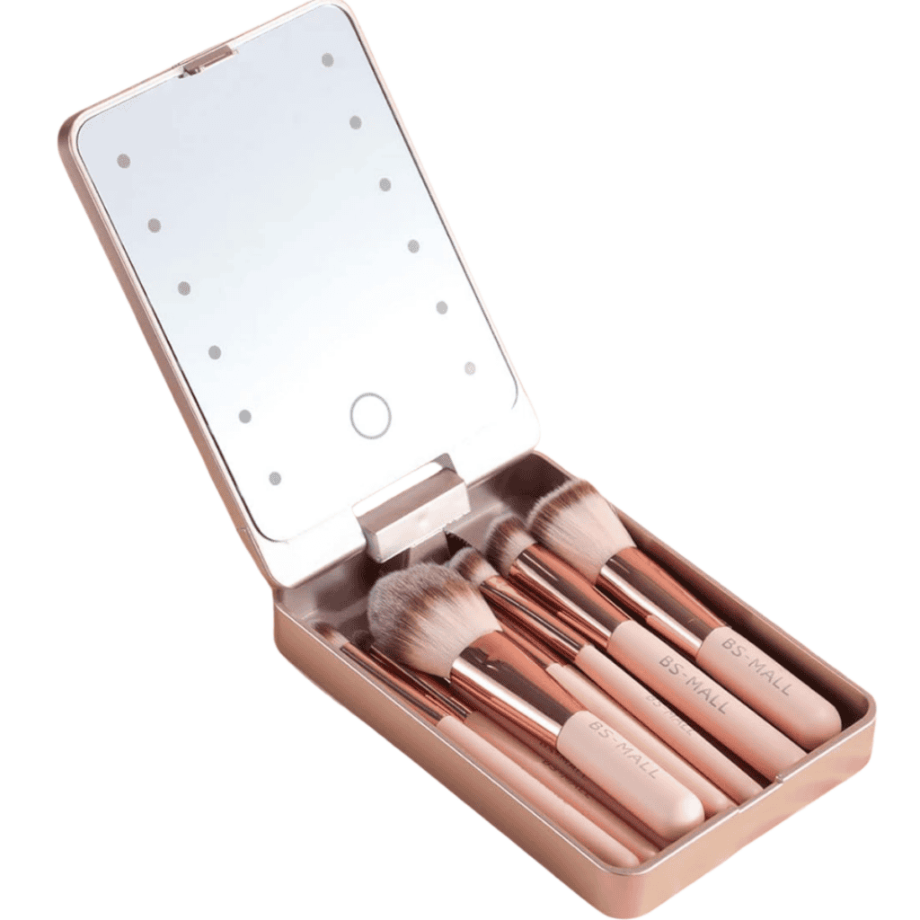 An open rectangular rose gold makeup case displays eight makeup brushes with rose gold handles and light brush heads. The lid features an illuminated mirror with LED lights around its border, a chic addition to any travel kit for those seeking elegant packing tips.